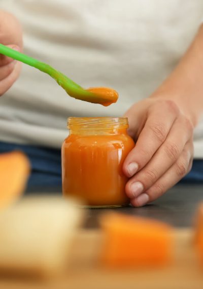 Close-up of a person scooping orange baby food from a jar with a green spoon, with fruits and toys in the background.