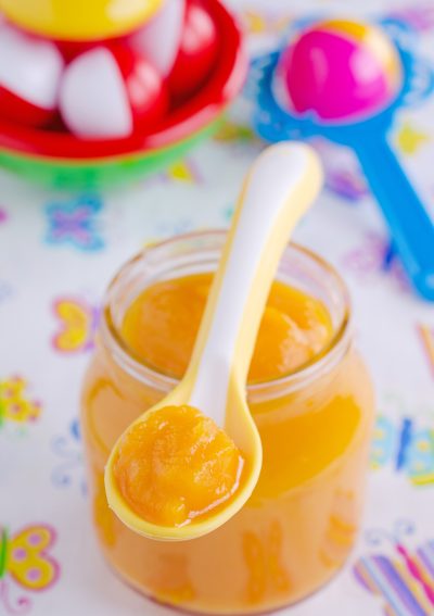 Glass jar of orange baby food puree with a spoon inside, placed on a colorful background.