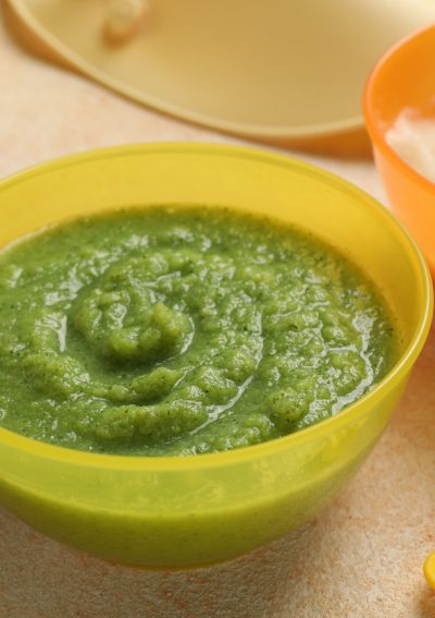 Green vegetable baby food puree in a bright green bowl on a kitchen counter. Next to an orange bowl with puree and baby spoons.