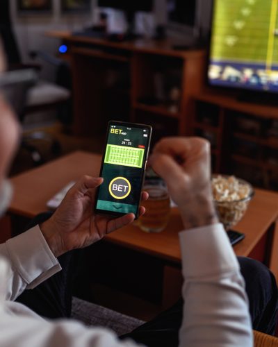 Man sitting on the sofa at home and watching soccer game. He is using smart phone for sports betting.