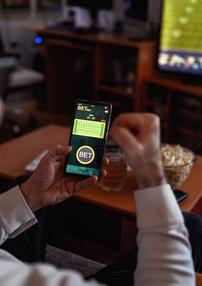Man sitting on the sofa at home and watching soccer game. He is using smart phone for sports betting.